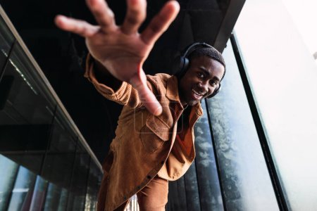 Téléchargez les photos : D'en haut d'homme afro-américain positif regardant la caméra et tendre la main tout en écoutant de la musique dans les écouteurs dans la rue - en image libre de droit