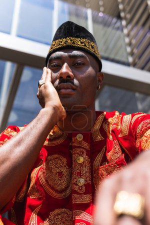 Ernsthafter erwachsener afrikanischer Typ in traditioneller roter Kleidung und Kufi-Mütze, der in die Kamera blickt, während er an sonnigen Tagen in der Stadtstraße in der Nähe eines Gebäudes mit Glaswänden sitzt