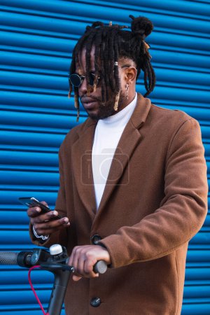 Photo for Trendy hipster African American guy in stylish coat and with dreadlocks browsing mobile phone while riding electric scooter against blue wall - Royalty Free Image