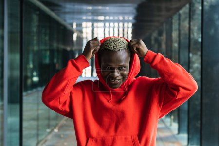Téléchargez les photos : Auto assuré jeune afro-américain millénial mâle avec capuche ajustable teint et regardant la caméra dans le passage moderne - en image libre de droit