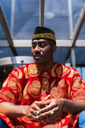 Ernsthafter erwachsener afrikanischer Typ in traditioneller roter Kleidung und Kufi-Mütze, der bei sonnigem Wetter in der Stadtstraße in der Nähe eines Gebäudes mit Glaswänden sitzt und wegschaut