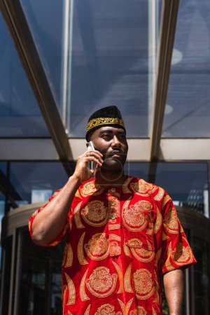 Hombre negro adulto serio en ropa roja tradicional y kufi de pie cerca del edificio con paredes de vidrio mientras conversaba por teléfono en un día soleado en la calle de la ciudad y mirando hacia otro lado