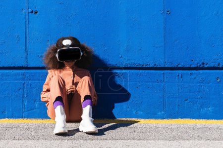 Unerkennbares afroamerikanisches Mädchen in schickem Anzug und VR-Headset erkundet virtuelle Welt, während sie auf der Straße vor blauer Wand sitzt
