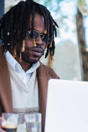 Téléchargez les photos : Homme afro-américain immoral assis à table avec un verre d'eau et expresso dans un café de rue et tapant sur un ordinateur portable - en image libre de droit