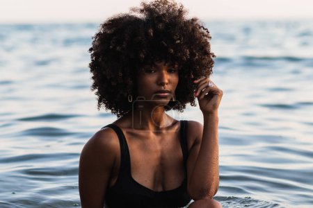 Photo for Tranquil black female wearing bikini standing in calm sea water in summer and looking away - Royalty Free Image