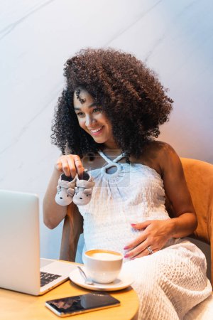 Foto de Positiva mujer afroamericana sentada en la mesa con taza de café con leche y mostrando botines de bebé a netbook durante la videollamada - Imagen libre de derechos