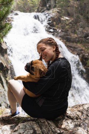 Delighted female hiker embracing French Bulldog near waterfall in woods while enjoying trekking in Pyrenees