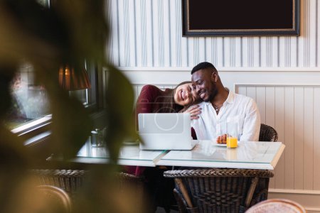 Téléchargez les photos : Heureux couple multiracial navigation ordinateur portable ensemble tout en étant assis à la table dans une chambre élégante à la maison - en image libre de droit