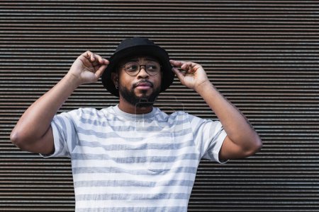 African American bearded guy with missing tooth in eyeglasses wearing trendy casual outfit and hat looking away while standing against shuttered wall