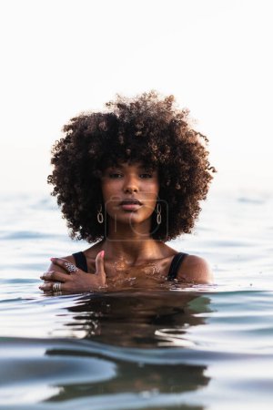 Calma hermosa mujer afroamericana en bikini y con peinado afro de pie en el agua del mar y mirando a la cámara