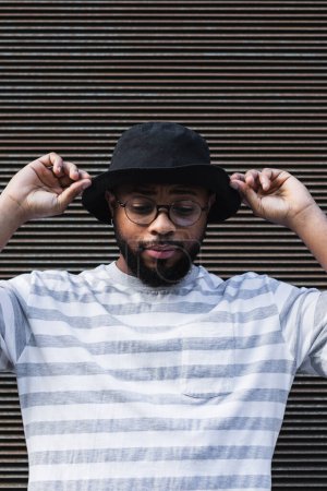African American bearded guy with missing tooth in eyeglasses wearing trendy casual outfit and hat looking down while standing against shuttered wall