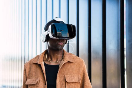 Afroamerikaner mit VR-Brille erkundet den Cyberspace, während er auf der Straße in der Nähe eines modernen Gebäudes mit Metallwand in der Stadt steht