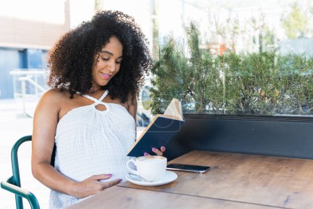 Photo for Young pregnant African American female with curly hair sitting at table and reading book while drinking coffee - Royalty Free Image