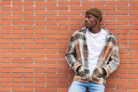 Téléchargez les photos : Jeune Afro-Américain en chemise à carreaux et chapeau à la mode fronçant les sourcils et détournant les yeux tout en se tenant contre le mur de briques sur la rue de la ville - en image libre de droit
