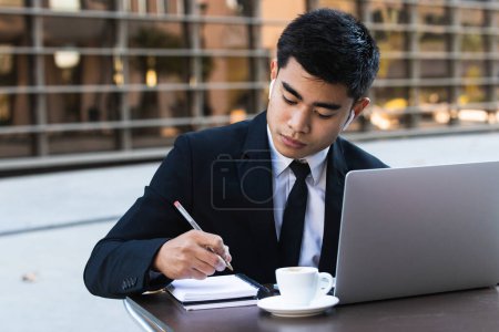 Téléchargez les photos : Focalisé homme d'affaires asiatique dans les écouteurs prendre des notes dans le bloc-notes tout en étant assis à table dans un café avec ordinateur portable et projet d'entreprise de planification - en image libre de droit