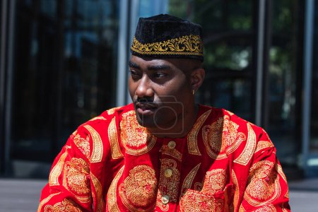 Chico africano adulto serio con ropa roja tradicional y gorra kufi mirando hacia otro lado mientras está sentado en la calle de la ciudad cerca del edificio con paredes de vidrio en un día soleado
