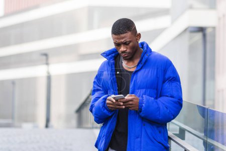 Hombre afroamericano con chaqueta abrigada parado en la calle cerca del edificio y mensajería en las redes sociales a través de teléfonos inteligentes