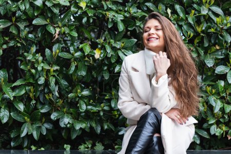 Photo for Charming elegant adult long haired brunette in stylish white coat smiling happily while resting near green bushes in park - Royalty Free Image