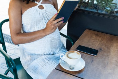 Téléchargez les photos : Cultivé femme afro-américaine enceinte méconnaissable avec les cheveux bouclés assis à la table et le livre de lecture tout en buvant du café - en image libre de droit