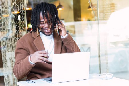 Téléchargez les photos : Homme noir hipster positif avec des tresses afro tenant un verre de boisson et parlant sur smartphone tout en étant assis à table avec un ordinateur portable et travaillant sur un projet à distance dans un restaurant extérieur - en image libre de droit