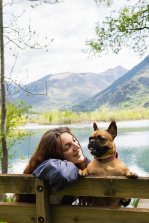 Foto de Mujer viajera encantadora con lindo Bulldog francés cerca del estanque en la cordillera de los Pirineos - Imagen libre de derechos