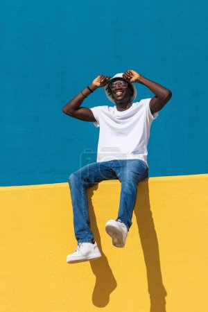 Foto de Low angle full body of optimistic young black male in casual outfit with panama hat and sunglasses smiling happily while siting on bright colorful fence and enjoying sunny summer day - Imagen libre de derechos