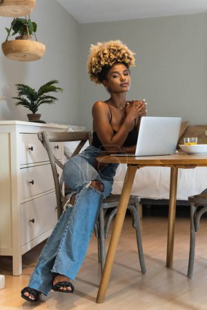 Téléchargez les photos : Corps complet de femme afro-américaine attrayante freelance assis regardant la caméra sur la chaise et regardant ordinateur portable à table avec petit déjeuner - en image libre de droit