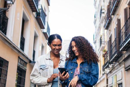 Téléchargez les photos : Deux amis latino-américains marchent dans la rue, s'amusent et rient en utilisant un smartphone. - en image libre de droit