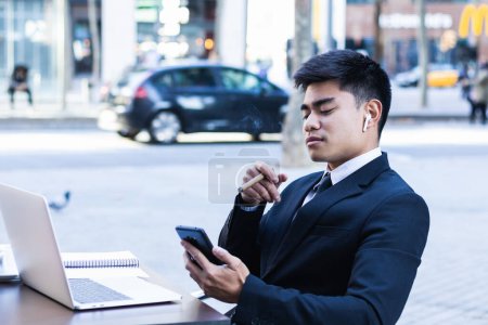 Téléchargez les photos : Confiant asiatique mâle entrepreneur assis à la table dans le café et fumer cigare tout en naviguant smartphone - en image libre de droit