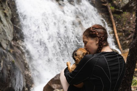 Delighted female hiker kissing French Bulldog near waterfall in woods while enjoying trekking in Pyrenees