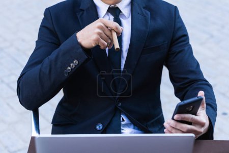 Photo for Confident Asian male entrepreneur sitting at table in cafe and smoking cigar while browsing smartphone - Royalty Free Image