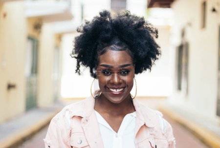 Portrait of a pretty black woman with afro hair looking at the camera. She has slightly spaced teeth and characteristic features. She looks happy.