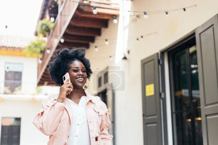 Elegante mujer negra con cabello afro caminando por la calle de la ciudad mientras habla por teléfono móvil y ríe. Lleva ropa casual y pendientes grandes..