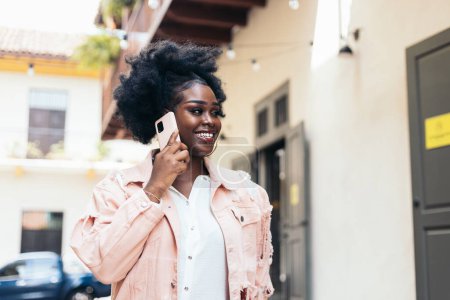 Femme noire élégante avec des cheveux afro marchant dans la rue de la ville tout en parlant sur le téléphone portable et en riant. Elle porte des vêtements décontractés et de grandes boucles d'oreilles.