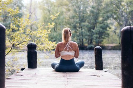 Téléchargez les photos : Jeune femme en vêtements de sport méditant et faisant du yoga dans la nature. Elle est pieds nus sur une plate-forme en bois près d'un lac - en image libre de droit