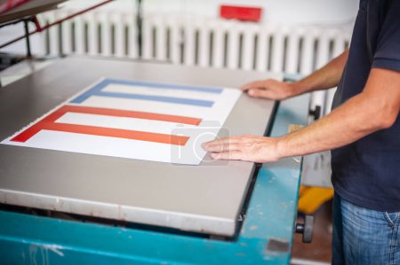 Photo for In the printing house, an experienced technician works on a screen printing machine. Production work. Check the print quality. - Royalty Free Image