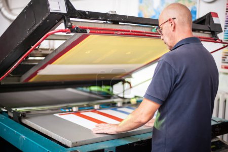 In der Druckerei arbeitet ein erfahrener Techniker an einer Siebdruckmaschine. Produktionsarbeit. Überprüfen Sie die Druckqualität.
