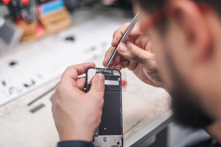 Blick von oben auf den Arbeitsplatz, Nahaufnahme. In einer Elektronik-Werkstatt repariert ein Reparateur ein Smartphone, benutzt eine Pinzette als eines der vielen Arbeitswerkzeuge.