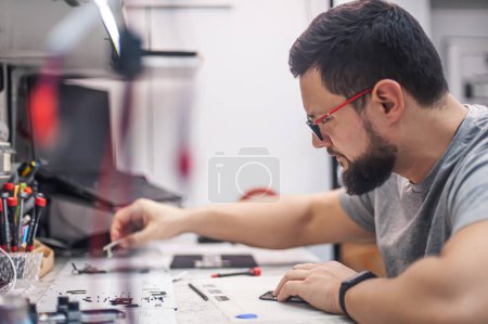 Electronics repair shop, a repairman is surrounded by tools and equipment. A technician repairs, cleans, controls a smartphone. Workplace top view, close-up.