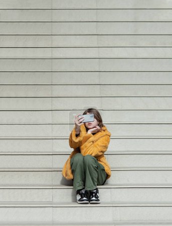 Téléchargez les photos : Jeune jolie femme en manteau de fourrure jaune, influenceuse des médias sociaux, assise sur les escaliers de la ville du bâtiment à l'aide d'un smartphone. - en image libre de droit