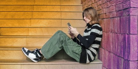 Téléchargez les photos : Jeune jolie femme dans un pull rayé noir et blanc, influenceur des médias sociaux, assis sur les escaliers de la ville du bâtiment en utilisant un smartphone. - en image libre de droit