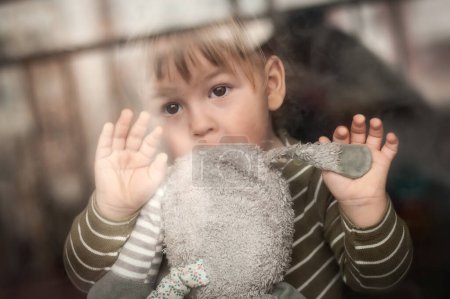 Téléchargez les photos : Portrait de mignon petit garçon avec jouet en peluche regardant par la fenêtre, regardant triste. Amour et émotion familiale - en image libre de droit