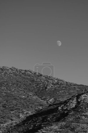 Photo for View of a mountain with a single white chapel and the moon in the blue sky in Ios Greece in black and white - Royalty Free Image
