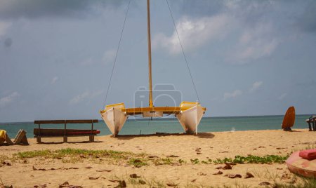 A serene image of a catamaran resting on a sandy beach with a calm sea and overcast sky in the background. The tranquil setting highlights the simplicity and beauty of coastal life.