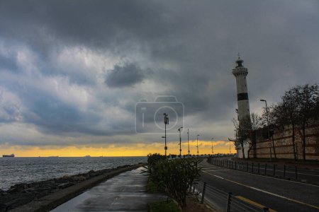 Lighthouse stands against a story sky over the road on a point with ships in the distance.