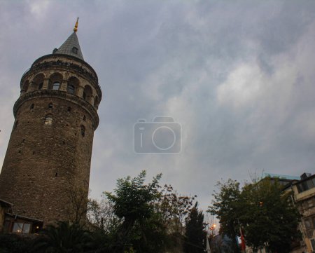 A captivating view of the historic Galata Tower in Istanbul, set against a cloudy sky. This ancient structure, with its distinctive conical roof, stands tall amidst modern buildings and lush greenery, showcasing the blend of history and urban life.
