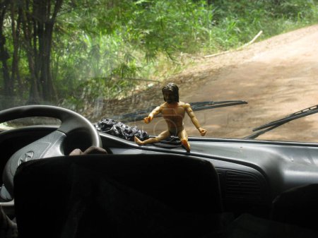 A whimsical shot of a Jesus action figure sitting on a car dashboard, with a forest road visible through the windshield. The scene captures a blend of playful and adventurous vibes.