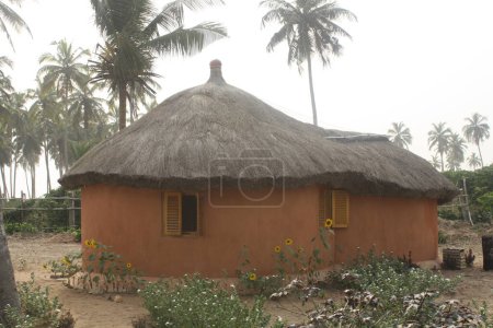 A traditional round hut with a thatched roof, set in a serene environment surrounded by palm trees. The scene reflects the simplicity and cultural heritage of rural life in Ghana.