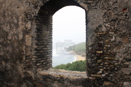 Ein gewölbter Fensterblick vom Fort Amsterdam, Ghana, mit Blick auf den weiten Ozean und die Küste, umrahmt von der rustikalen Backsteinstruktur des Forts