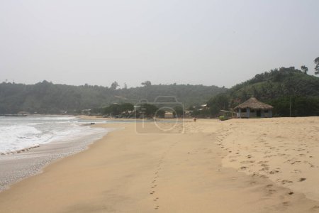 Ein ausgedehnter Strand mit sanften Wellen, die an Land spülen und Fußspuren entlang des Sandstrandes, mit üppigem Grün im Hintergrund.